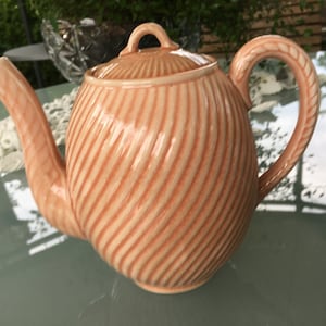 Può includere: Una teiera in ceramica color pesca con un design a spirale e nervature. La teiera ha un coperchio e un manico.