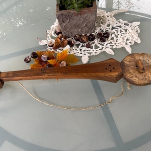Puede incluir: Un instrumento musical de madera con un cuerpo redondo y desgastado y un cuello largo tallado. El instrumento está encordado con una sola cuerda y tiene un pequeño puente de madera. El instrumento está sobre una mesa con un mantel blanco y algunas hojas de otoño.