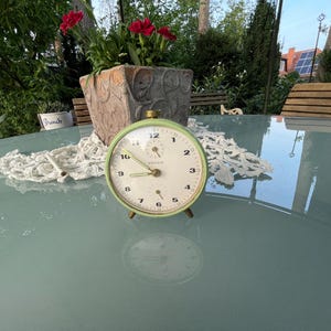 Könnte beinhalten: Ein grüner Vintage-Wecker mit weißem Zifferblatt und schwarzen Zahlen. Der Wecker steht auf einem hellblauen Tisch mit einer weißen Spitzendeckchen.