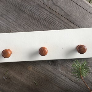 Może przedstawiać: Biały drewniany wieszak na ubrania z trzema brązowymi drewnianymi gałkami.