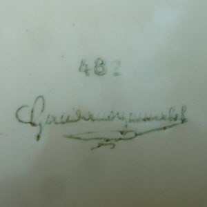 May include: A close-up of a white ceramic plate with a green handwritten signature that reads "Friedrich  H" and the number "482" written in green above the signature.