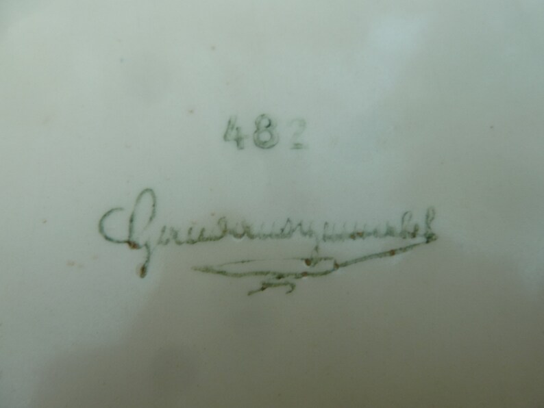 May include: A close-up of a white ceramic plate with a green handwritten signature that reads "Friedrich  H" and the number "482" written in green above the signature.