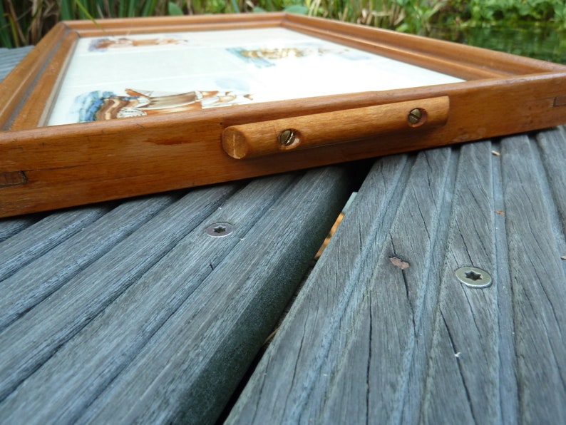 May include: A wooden tray with a white tile insert and a brass handle. The tray is resting on a gray wooden surface.