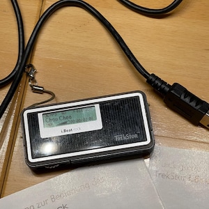 Peut inclure: Lecteur MP3 TrekStor i.Beat rock noir et argenté avec un petit écran affichant "Choo Choo" et "i.Beat rock". Un câble USB noir est connecté à l'appareil. L'appareil est sur une surface en bois.