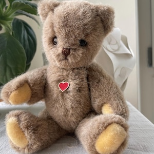 Puede incluir: Un oso de peluche marrón con un colgante de corazón rojo. El oso de peluche tiene una textura suave y peluda, con patas de color canela y una nariz y ojos pequeños y oscuros. El oso está sentado erguido, con los brazos y las piernas extendidos.