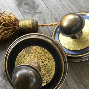 Könnte beinhalten: Zwei goldfarbene Metall-Vorhangbinder mit schwarzen Akzenten und einem goldenen Blumenmuster. Jeder Vorhangbinder hat einen runden Knopf und einen Quasten.