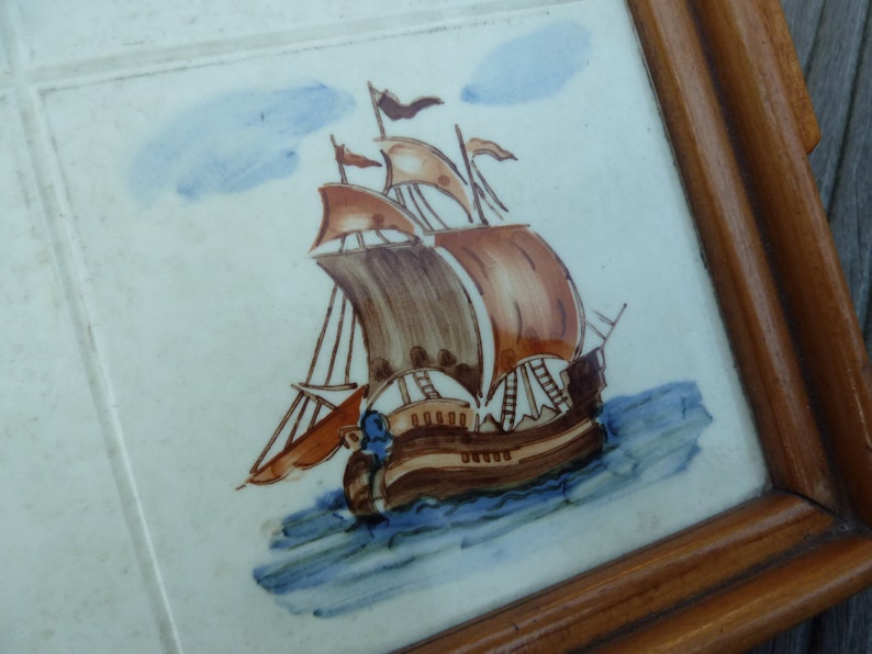 May include: A framed tile with a hand-painted image of a brown and white sailing ship with blue and white waves. The ship has brown sails and a blue and white flag.