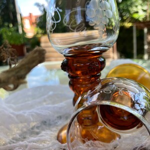 Könnte beinhalten: Drei Weingläser mit Stiel aus bernsteinfarbenem Glas mit gravierten Traubenmotiven. Die Gläser sind übereinander gestapelt.
