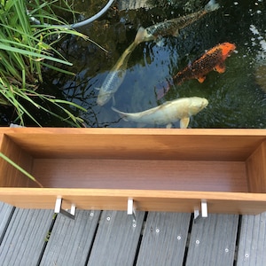 Könnte beinhalten: Ein Holz-Pflanzgefäß mit einer natürlichen Holzoptik und silbernen Metallhalterungen. Der Pflanzkasten ist leer und steht auf einem Holzdeck.