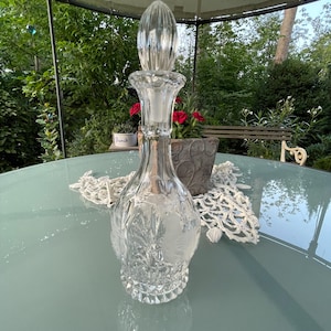Peut inclure: Une carafe en verre transparent avec une surface texturée et un bouchon rond en verre transparent. La carafe est posée sur une nappe de table blanche avec une table bleu clair en dessous.