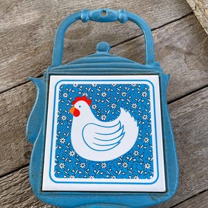 May include: Blue cast iron teapot hot pad with a white ceramic tile featuring a white chicken with a red comb and wattle on a blue background with white flowers.