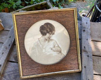 May include: A framed sepia print depicting a mother and baby. The artwork is set within a rectangular wooden frame with a gold-coloured inner border. The image is a vintage portrait, likely from the early 20th century.