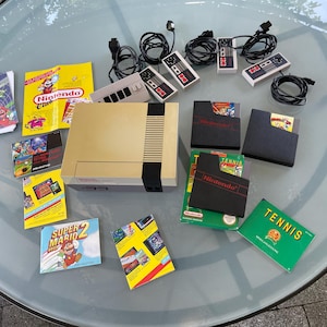 Op de afbeelding: Een beige Nintendo Entertainment System (NES) console met controllers, spelcartridges en handleidingen. Spellen zijn onder andere Super Mario 2 en Tennis. De controllers en de console zijn zwart en grijs.