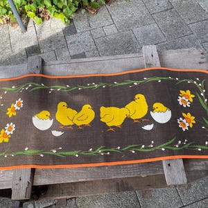 Op de afbeelding: Een rechthoekige bruine mat met een oranje rand, met gele kuikens, witte eierschalen en bloemmotieven. Het ontwerp bevat groene wijnranken en gele narcissen, wat een lente- of Paas thema creëert.