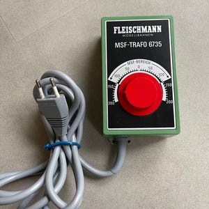 May include: A green and black Fleischmann MSF-TRAFO 6735 model railway transformer with a red dial. The dial has white markings from 0 to 200. A grey power cord with a two-pin plug is connected to the transformer.