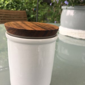 Pode incluir: Um pote de cerâmica branco com uma tampa de madeira. O pote está sobre uma mesa de vidro.