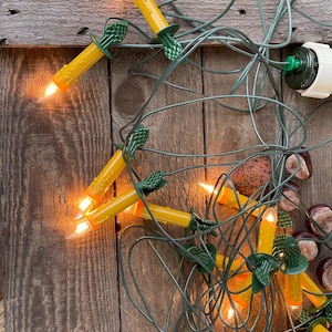 Op de afbeelding: Een reeks vintage kerstverlichting met gele kaarsvormige lampen en groene bedrading. De lichten liggen op een verweerd houten oppervlak, met enkele kastanjes. De lampen zijn verlicht en geven een warme, uitnodigende gloed.