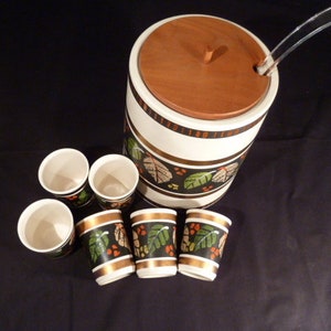 Può includere: Una ciotola per punch in ceramica bianca con un coperchio in legno marrone e una paletta in plastica trasparente. La ciotola ha una fascia nera e dorata con un motivo a foglie verdi e arancioni. Sono incluse sei tazze in ceramica abbinate con lo stesso motivo.