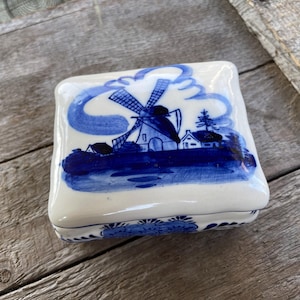 Könnte beinhalten: Eine weiße Keramik-Schmuckschatulle mit einer blauen und weißen Windmühlen-Szene. Die Schachtel hat eine quadratische Form und eine erhabene Verzierung an den Rändern.