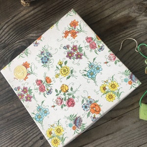 Può includere: Una scatola regalo bianca con un motivo floreale in tonalità di rosa, giallo, blu e verde. La scatola è decorata con una moneta d'oro. Un ciondolo a forma di farfalla verde e oro pende da un cordoncino verde.