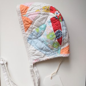 May include: A white baby bonnet with a patchwork design featuring various colors and patterns, including pink, blue, red, and orange. The bonnet has a white tie and a tag that reads "Cotton Pie".