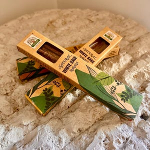 May include: Several boxes of natural white sage incense sticks. The boxes are rectangular with a brown and green design, and the text "Natural White Sage Masala" is printed on the side. The incense sticks are visible through a rectangular opening.