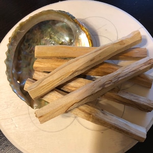 Può includere: Un set di sei bastoncini di Palo Santo in legno disposti su un vassoio circolare in legno con una grande conchiglia di abalone. I bastoncini sono di colore marrone chiaro e hanno una texture naturale e ruvida.