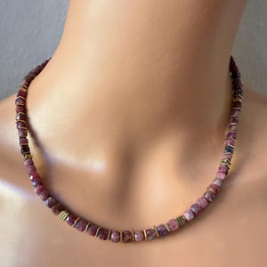 Könnte beinhalten: Eine Halskette mit facettierten, rot-violetten Edelsteinen, die von kleinen goldfarbenen Abstandshaltern unterbrochen werden. Die Halskette wird am Hals und der Brust einer Person präsentiert und zeigt das Design und die Farbe des Schmucks.