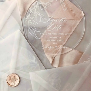 Könnte beinhalten: Eine transparente Acryl-Save-the-Date-Karte mit weißen Blumenmustern und dem Text "Save the Date" in einer Schreibschrift. Der Text "Do all the things with love" befindet sich unter dem Datum. Das Datum ist "Deidesheim, Sonntag, 7. März 2021". Die Namen "Carolyn & John" stehen unten auf der Karte.