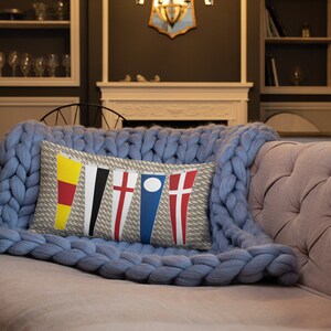 May include: A decorative pillow with a nautical theme featuring five colorful pennants with rope accents. The pennants are yellow, black, red and white, blue and white, and red and white. The pillow is on a blue knitted throw blanket.