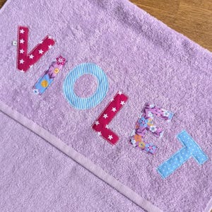 Könnte beinhalten: Ein hellviolettes Frotteehandtuch mit dem Namen VIOLET, der in verschiedenen Stoffmustern appliziert ist. Der Buchstabe V ist dunkelrosa mit weißen Sternen, I ist floral, O ist hellblau gestreift, L ist dunkelrosa mit weißen Sternen, E ist floral und T ist hellblau mit weißen Punkten.