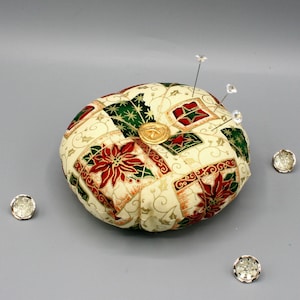 May include: A round pincushion with a cream background and a patchwork design featuring red poinsettia flowers, green leaves, and gold accents. The pincushion has three decorative pins with clear crystal tops.