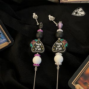 May include: Two decorative pins with silver-colored shafts. Each pin features a black bead, a white bead with a purple band, and a black charm with the text "Fuck all y'all". The pins are topped with a small skull and a hand holding a planchette.