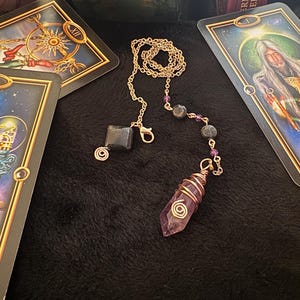 May include: A gold-colored chain with a faceted black stone and a purple amethyst crystal pendulum. The amethyst is wrapped in copper wire. Tarot cards are in the background.