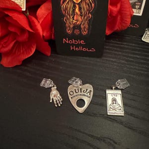 May include: A collection of tarot cards, red roses, and three silver charms. The charms include a hand, a heart-shaped Ouija planchette, and a rectangular pendant with a woman's image. The text "Noble Hollow" is visible on a card.