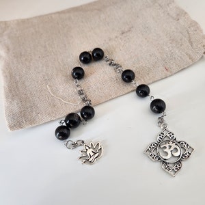 May include: Black beaded mala with silver Om and lotus charms. The mala is in a small beige drawstring bag.
