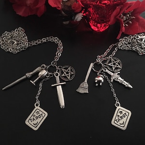 May include: Two silver-toned necklaces with various charms, including a pentagram, sword, broom, and tarot card. One necklace features a wand and chalice charm. The tarot card charm reads "TAROT". The necklaces are displayed on a black surface.