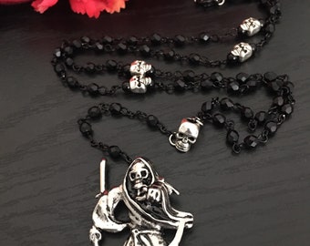 Puede incluir: Un collar de cuentas negras con detalles de calaveras y un colgante plateado de la Parca. El collar se exhibe sobre una superficie oscura, con flores rojas y una decoración en forma de calavera en el fondo. Aproximadamente 45 cm de largo.