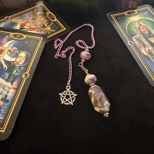 Könnte beinhalten: Ein lila Kettenpendel mit einem silbernen Pentagramm-Anhänger und Amethystperlen. Das Pendel wird mit Tarotkarten auf einer schwarzen Oberfläche präsentiert. Die Karten haben goldene Akzente und zeigen Illustrationen.