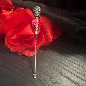 May include: A decorative stick pin featuring a metallic silver skull topper, a clear glass bead containing a pink rose, and small black accent beads. The pin has a long, thin silver-toned metal shaft that tapers to a point with a clear plastic cap.