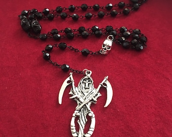 Puede incluir: Collar de rosario de cuentas negras con un colgante de plata que representa una figura esquelética sosteniendo guadañas.
