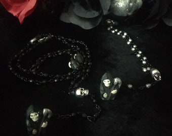 Puede incluir: Un collar de cuentas negras con tres dijes plateados, dos de los cuales son Parcas y uno es una calavera.