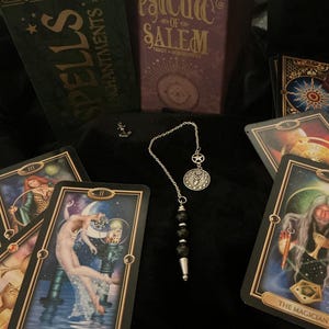 Pode incluir: Uma composição de cartas de tarot, livros e um pêndulo. Os livros são intitulados "Spells & Enchantments" e "Psychic of Salem". O pêndulo tem uma corrente prateada, um berloque de estrela e um pingente circular. As cartas de tarot apresentam ilustrações.