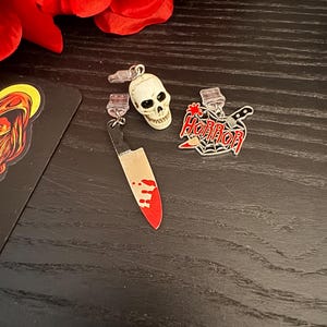 Puede incluir: Una colección de pendientes con temática de Halloween. Un pendiente presenta una pequeña calavera blanca, otro un cuchillo con una hoja rojo sangre, y el tercero tiene la palabra "HORROR" con una telaraña y un cuchillo. Rosas rojas en el fondo.