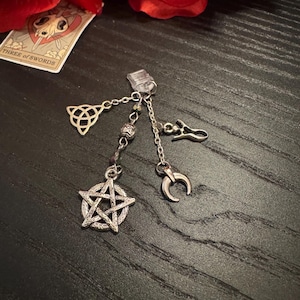 May include: Silver charm with a clear clip, featuring a pentagram, crescent moon, Celtic knot, and other charms. The charms are connected by delicate chains and beads. A tarot card and red flowers are in the background.