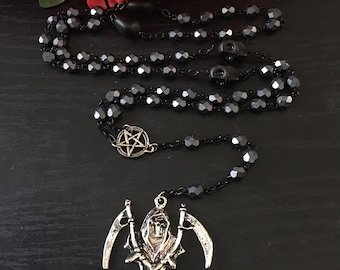 Puede incluir: Collar de rosario de cuentas negras con un colgante de plata que representa una figura esquelética sosteniendo dos guadañas. El colgante está unido a una cadena negra con un amuleto de pentagrama.