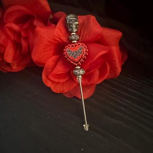 May include: A decorative stick pin featuring a metallic skull topper, two textured silver-toned beads, and a central red heart-shaped charm with a black rose pattern. The pin has a long, thin metal shaft and a small clear plastic end cap.