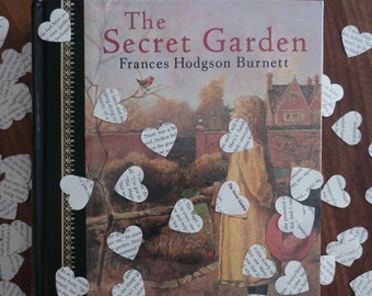 May include: A vintage copy of "The Secret Garden" by Frances Hodgson Burnett, illustrated in color by Troy Howell. The book cover features a colorful illustration of a girl in a yellow coat standing in a garden with a robin perched on a branch above her. The book is surrounded by white paper hearts with text printed on them.