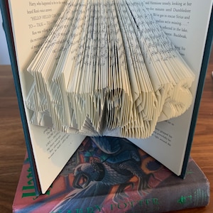 May include: A book with pages folded into a 3D shape resembling a bird. The book is titled "Harry Potter and the Prisoner of Azkaban" and is sitting on a wooden surface.