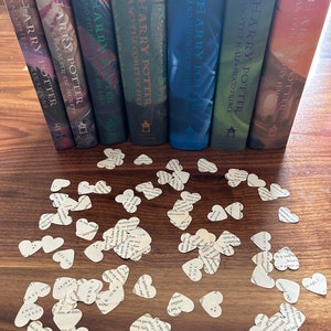 May include: A set of seven Harry Potter books, with the spines facing out, are arranged in order from left to right. The books are on a wooden table with a scattering of paper heart confetti.